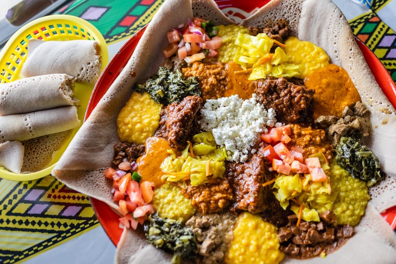 Colorful Ethiopian injera platter with various stews, vegetables, and cheese, with rolled injera on the side.
