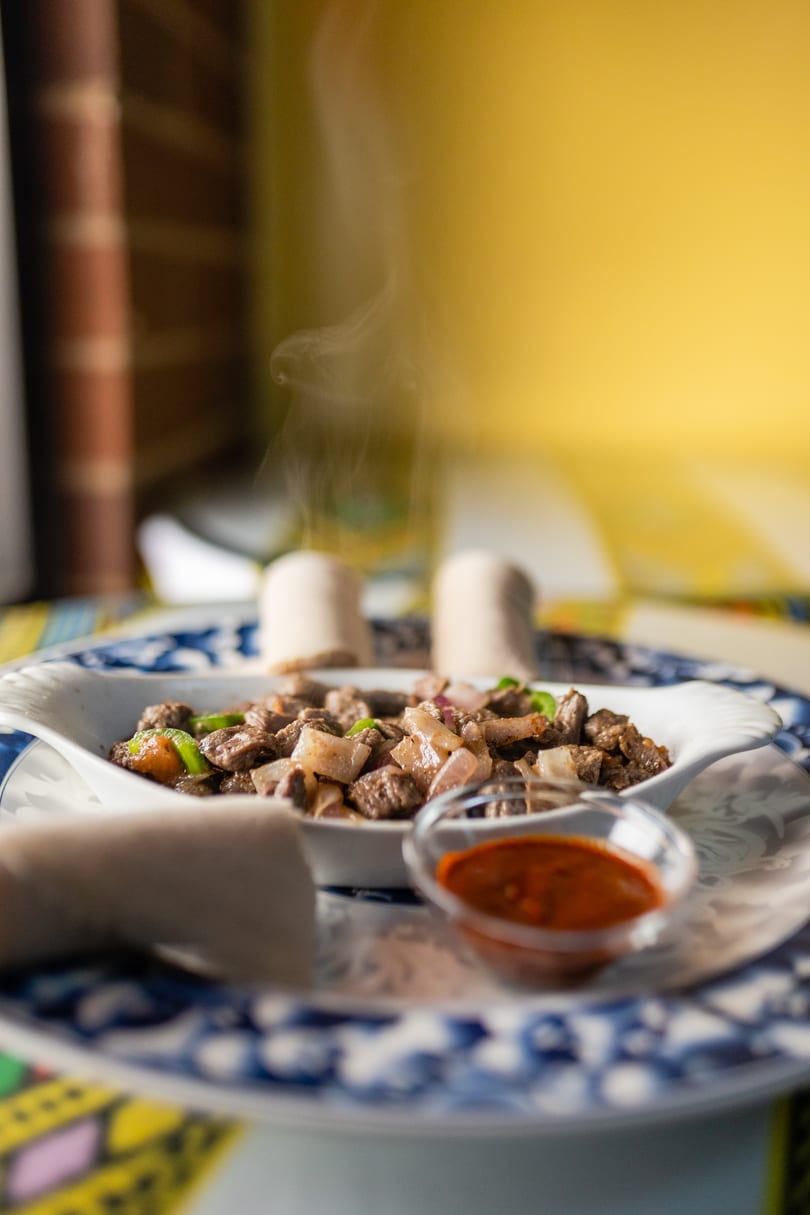 Steaming hot Ethiopian tibs with injera bread and a spicy dipping sauce on a patterned plate.