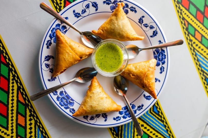 Five golden-brown samosas and a bowl of green chutney on a white plate with blue floral designs.