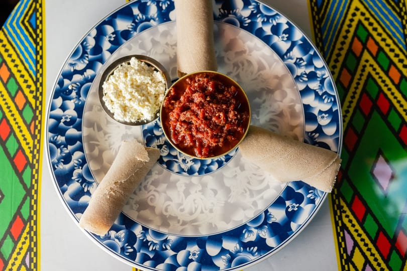 Overhead view of injera rolls, red stew, and white cheese on a blue and white plate.