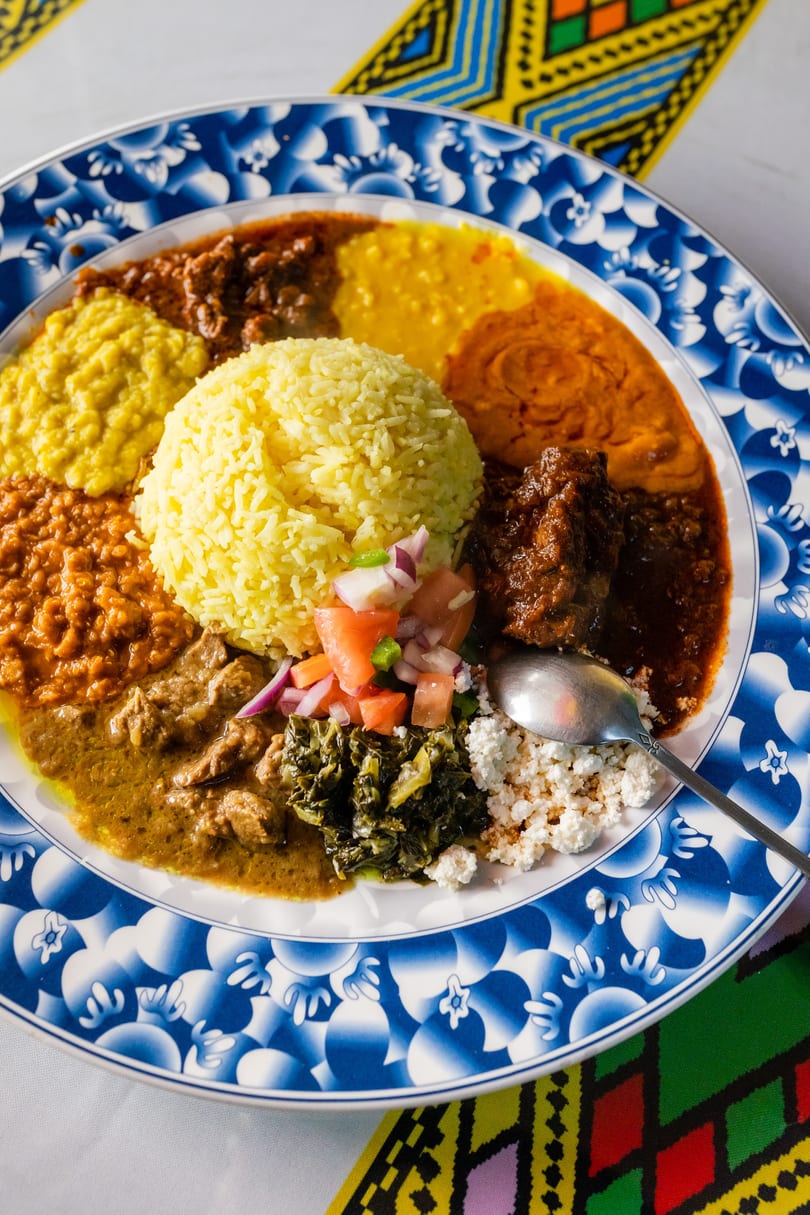 A colorful Ethiopian platter with yellow rice, various stews, vegetables, and cheese on a blue and white plate.